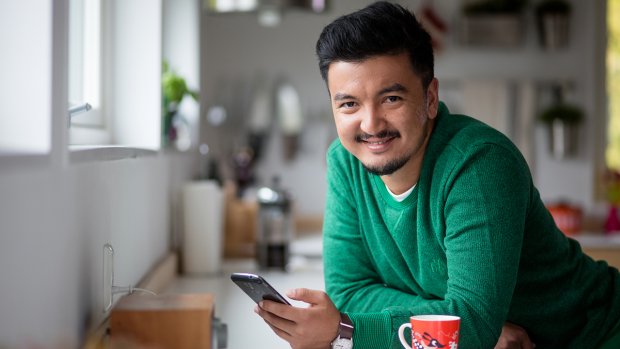 Mann står ved kjøkkenbenken og holder en mobil. Ser i kameraet og smiler.