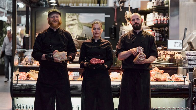 To menn og en kvinne i svarte kokkeklær med matprodukter foran slakterbutikk